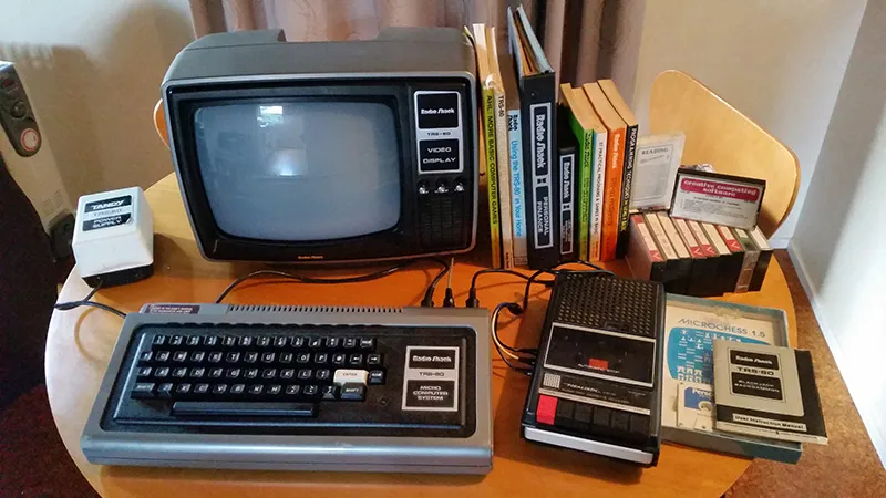 A TRS-80 sits with monitor and tape deck drive sits on a small kitchen table next to manuals and cassettes. In the background is a space heater and curtains